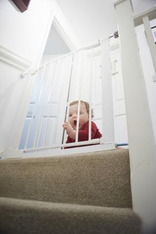 Cancello di sicurezza per bambini sulle scale: bambino sul pianerottolo di casa sua. È dietro un cancello di sicurezza per bambini che lo impedisce dal pericolo della scala.