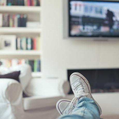 Immagine di jeans e scarpe da ginnastica, i piedi sul divano, con televisione, camino, divano e libreria in background.